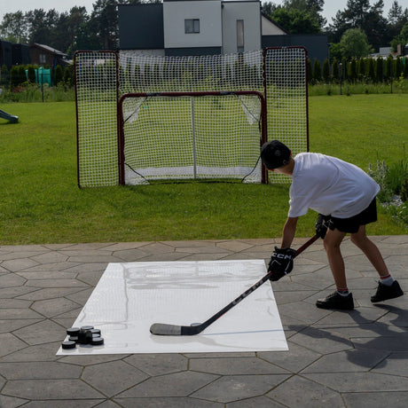 Young hockey player practicing shots on a roll-up shooting pad in a garden with a full-size goal